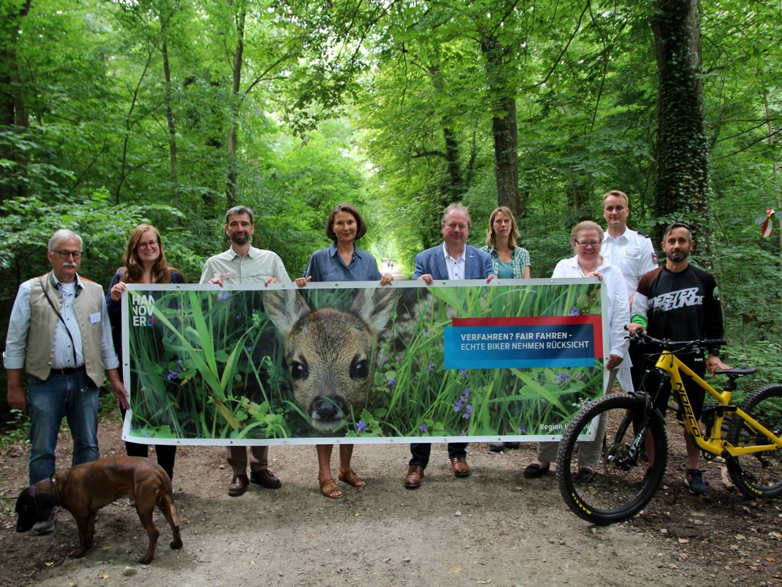 Eine Gruppe von Menschen steht auf einem Waldweg und hält ein Banner in den Händen - flankiert von einem Mountainbike und einem süßen Hund